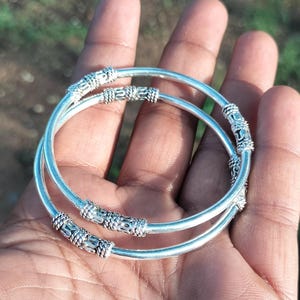 Puede incluir: Dos pulseras de plata con secciones decorativas de cuentas. Las pulseras son redondas y lisas, con patrones detallados en las secciones de cuentas. Las joyas se muestran en una mano, con un fondo verde borroso.