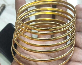 Könnte beinhalten: Ein Stapel von sieben goldfarbenen Armreifen, jeder mit einer glatten, abgerundeten Oberfläche. Die Armreifen werden in der Hand einer Person gehalten, wobei sich das Licht auf dem polierten Metall spiegelt. Der Schmuck scheint aus einem festen Material zu bestehen.