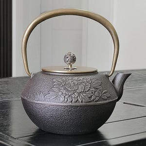 Puede incluir: Tetera de hierro fundido negro con diseño floral y asa de latón. La tapa es dorada y está rematada con un pomo decorativo. La tetera está sobre una superficie negra.