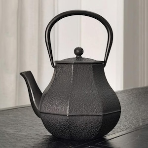 May include: A black cast iron teapot with a textured surface. The teapot has a curved handle, a spout, and a lid with a small knob. The teapot is sitting on a dark surface.