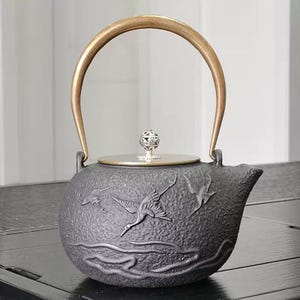 Puede incluir: Una tetera de hierro fundido negro con asa y tapa doradas. La tetera presenta un diseño en relieve de grullas en vuelo. La tapa está rematada con un pomo decorativo plateado. La tetera está diseñada para preparar té.