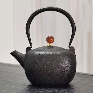 Puede incluir: Tetera de hierro fundido negro con superficie texturizada, asa curva y boquilla pequeña. La tapa tiene un pomo decorativo con base dorada y esfera marrón rojiza. Diseñada para preparar té.