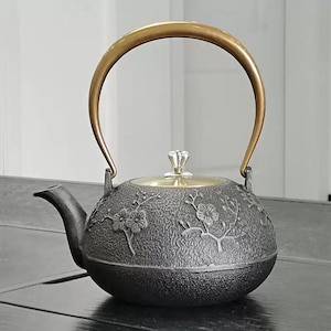 Puede incluir: Tetera de hierro fundido de color gris oscuro con diseños florales en relieve. Tiene un asa y una tapa doradas. El pico de la tetera es curvo y el asa forma un arco en la parte superior.