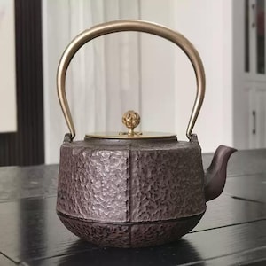 Puede incluir: Una tetera de hierro fundido marrón oscuro con una superficie texturizada, un asa dorada y una tapa a juego. La tetera tiene un pico y un pomo decorativo en la tapa. El asa se arquea.