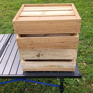 May include: A rectangular wooden crate constructed from light-colored horizontal planks. The crate features a solid top surface made of parallel wood slats and open gaps between the side panels, held together by visible metal screws at the corners.