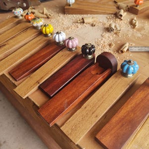 May include: Handmade wooden doorstops with colourful ceramic knobs. The doorstops are made from different types of wood and are displayed on a wooden surface. The knobs are in various colours, including orange, yellow, pink, and blue, showcasing craftsmanship.