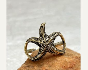 May include: A gold-toned metal ring featuring a textured starfish design. The starfish has a detailed, bumpy surface with dark oxidation in the crevices, creating a contrast against the metallic gold finish of the band and the starfish shape.