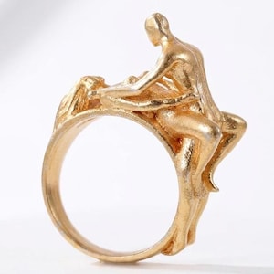 Puede incluir: Un anillo dorado con un diseño esculpido de dos figuras abrazándose. La banda del anillo es lisa, mientras que las figuras tienen detalles texturizados. El diseño general es artístico y detallado, con un enfoque en la forma humana.