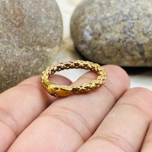 May include: A gold-coloured ring in the shape of a snake, with the head of the snake facing outwards. The ring has a textured, scale-like pattern. The ring is on a person's hand.