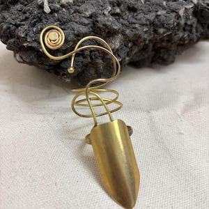 May include: A brass finger armor ring with a spiral design and a pointed end. The ring has a gold finish and is set against a dark, textured background. The ring's design includes a spiral and a small ball.