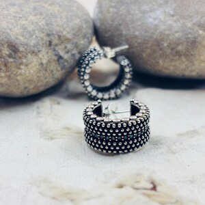 May include: Close-up of two silver hoop earrings with a textured, dotted design. The earrings are set against a neutral background with two large gray stones. The earrings have a classic, yet detailed design.