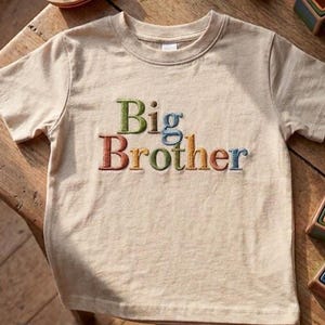 Op de afbeelding: Een beige t-shirt met de geborduurde tekst "Big Brother" in kleurrijke letters. Houten speelgoed, waaronder een boot en blokken, liggen verspreid rond het shirt op een houten oppervlak.