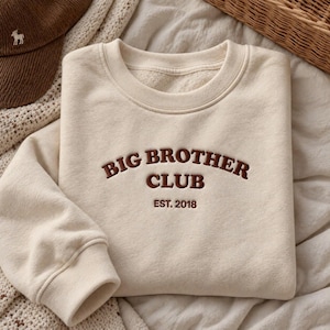 May include: A cream-colored crewneck sweatshirt featuring dark brown embroidered text that reads BIG BROTHER CLUB EST. 2018. The garment has long sleeves with ribbed cuffs and a ribbed neckline, showcasing a soft, fleece-lined interior texture.