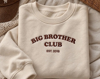 Può includere: Una felpa girocollo color crema con testo ricamato in marrone scuro che recita BIG BROTHER CLUB ed EST. 2018. Il capo presenta maniche lunghe, un colletto a coste e una morbida fodera interna in pile visibile sullo scollo.