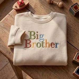 Könnte beinhalten: Cremefarbenes Sweatshirt mit dem gestickten Text "Big Brother" in bunten Buchstaben. Das Sweatshirt liegt gefaltet auf einer Holzoberfläche, mit einem kleinen Holzboot und anderem Spielzeug in der Nähe.