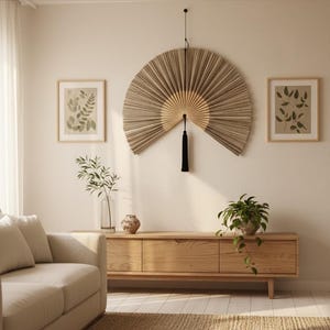 Puede incluir: Un abanico de pared beige con una borla negra cuelga sobre un mueble de madera. Dos estampados botánicos enmarcados y un sofá también están en la habitación. Una planta en maceta y un jarrón con vegetación se suman a la decoración. La habitación tiene una paleta de colores neutros.