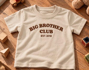 Op de afbeelding: Een beige t-shirt met korte mouwen en de bruin geborduurde tekst BIG BROTHER CLUB EST. 2018 in het midden op de borst. Het shirt heeft een ronde hals en is gemaakt van een effen stof met een eenvoudig en strak ontwerp.