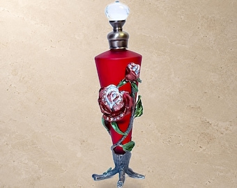 May include: A tall, conical red glass perfume bottle featuring a metallic rose embellishment with clear crystals on the front. The bottle has a silver-toned metal base shaped like tree roots and a clear, spherical glass stopper on top.