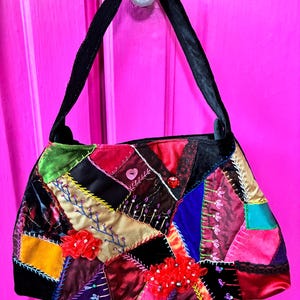 May include: A handbag featuring a patchwork design of various fabrics including velvet, satin, and ribbon in shades of red, black, gold, blue, and green. The bag is decorated with embroidery, small red fabric flowers, a pink heart button, and a red bead.
