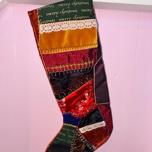 May include: A patchwork Christmas stocking featuring a dark green top band with the repeating text merry christmas, accented by white lace trim, gold velvet, deep red fabric, black floral patterns, and blue velvet sections with decorative stitching.