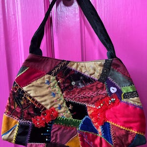 May include: A handbag featuring a patchwork design of various fabrics including velvet, satin, and corduroy in shades of red, gold, blue, and green. The surface is decorated with intricate embroidery, beads, sequins, and fabric flowers, with a black handle.