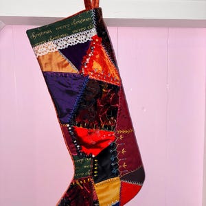 Op de afbeelding: Een patchwork kerstkous met een donkergroene boord met de herhaalde tekst "merry christmas" en witte kanten afwerking. Het lichaam bestaat uit diverse lapjes stof in rood, paars, oranje, zwart en goud, versierd met decoratieve stiksels en kralen.