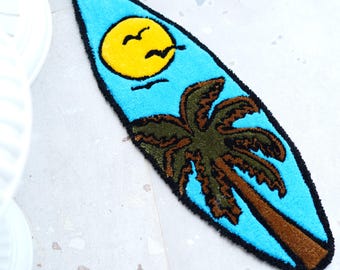May include: A blue surfboard-shaped rug featuring a yellow sun, birds, and a palm tree design. The rug has a black border and is made of a soft, textured material. The rug is designed for use in a bathroom or bedroom.