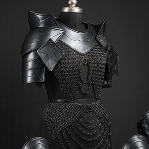 Puede incluir: Un conjunto de armadura negra que incluye una pieza de pecho con cota de malla integrada, hombreras y cuatro grebas a juego. La placa pectoral muestra un dise&ntilde;o en capas con cadenas colgantes, mientras que las grebas presentan una superficie lisa y curva con detalles en forma de V y remaches visibles.