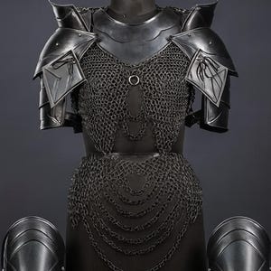 Puede incluir: Una armadura de metal oscuro que incluye un corpi&ntilde;o de cota de malla con cadenas drapeadas, hombreras s&oacute;lidas y cuatro grebas o brazales a juego expuestos en soportes individuales. El conjunto tiene un acabado en tono gris carb&oacute;n mate.