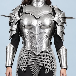 May include: A full set of polished silver-colored metal plate armor featuring a breastplate, articulated shoulder pauldrons, arm bracers, and greaves. The armor includes a chainmail skirt and is worn over a black long-sleeved shirt and black pants.