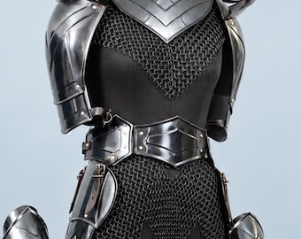 May include: A set of polished steel plate armor featuring a breastplate, shoulder pauldrons, arm guards, and leg greaves layered over a black chainmail tunic. The armor pieces are secured with dark leather straps and buckles on a display mannequin.