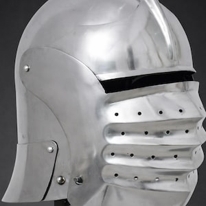 May include: A polished steel sallet helmet featuring a rounded skull cap, a prominent central ridge, and a hinged visor with horizontal ridges and ventilation holes. The helmet includes a black leather chin strap with a metal buckle attachment.