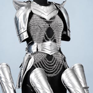 Op de afbeelding: Een set gepolijst zilverkleurig metalen harnas met een borstplaat met geïntegreerd maliënkolder, schouderstukken, een heupgordel en drie afzonderlijke scheenplaten voor beenbescherming. Het harnas heeft overal een gladde, reflecterende afwerking.