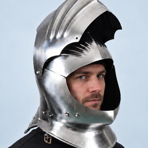 May include: A polished steel knight helmet featuring a raised visor, a rounded crown with layered metal plates, and a wide neck guard. The helmet is constructed from reflective silver-toned metal with visible rivets along the lower edge.