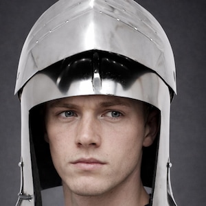 May include: A polished silver-colored metal bascinet helmet featuring a pointed visor and a rounded skull cap. The helmet includes a hinged visor mechanism with visible rivets along the edges and black leather chin straps hanging down on both sides.