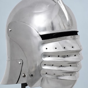 May include: A polished silver-colored metal helmet featuring a rounded dome, a prominent central ridge, and a hinged visor with horizontal ridges and multiple ventilation holes. The helmet includes black leather chin straps with metal buckles.