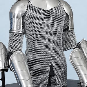 Könnte beinhalten: Eine silberne Metallrüstung bestehend aus einem Kettenhemd mit langen Ärmeln, einem Halsschutz, Schulterplatten und vier separaten Beinschienen oder Armschienen auf Ständern. Die Rüstungsteile haben eine polierte Oberfläche und bewegliche Platten.