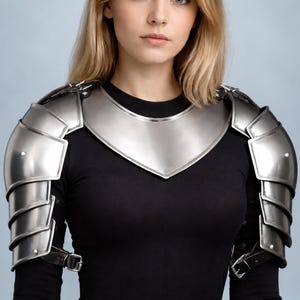 Op de afbeelding: Een set gepolijste stalen schouderstukken en een halskraag gedragen over een zwart shirt met lange mouwen. De schouderharnas bestaat uit overlappende metalen platen die zijn vastgezet met leren riemen en gespen, wat de schouders en de bovenborst bedekt.
