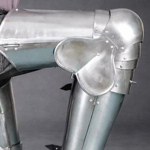May include: A set of polished steel leg armor featuring articulated thigh plates, knee cops with side wings, and greaves. The armor pieces are secured with black leather straps and metal buckles, covering the upper and lower legs in a metallic finish.