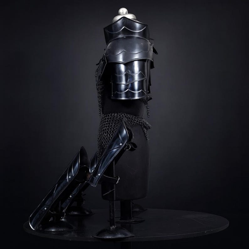 Puede incluir: Una armadura de metal negro con acabado pulido, que incluye un gorjal, hombreras articuladas y una cota de malla. El conjunto incluye grebas de metal negro a juego colocadas sobre soportes frente a la pieza del torso.