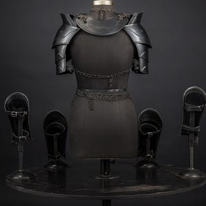 Puede incluir: Un conjunto de armadura de metal negro que incluye hombreras, un gorjal y detalles de cota de malla sobre un maniqu&iacute; de torso. Cuatro grebas o protectores de brazo de metal negro se exhiben en soportes alrededor de la pieza central.