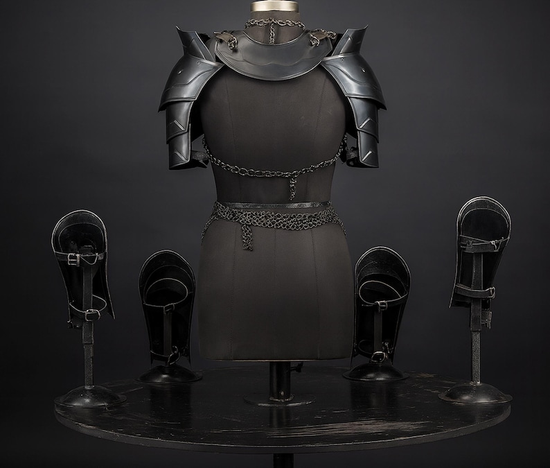 Puede incluir: Un conjunto de armadura de metal negro que incluye hombreras, un gorjal y detalles de cota de malla sobre un maniqu&iacute; de torso. Cuatro grebas o protectores de brazo de metal negro se exhiben en soportes alrededor de la pieza central.