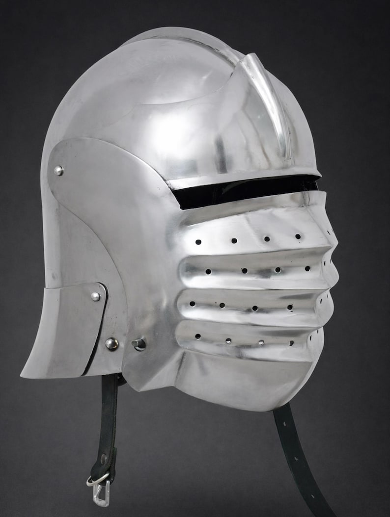 May include: A polished steel sallet helmet featuring a rounded skull cap, a prominent central ridge, and a hinged visor with horizontal ridges and ventilation holes. The helmet includes a black leather chin strap with a metal buckle attachment.