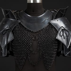 Puede incluir: Una armadura de pecho de metal negro que incluye un gorjal, hombreras articuladas con cordones negros y una cota de malla. La armadura presenta eslabones de cadena decorativos que cuelgan en la parte frontal, asegurados con remaches y placas.