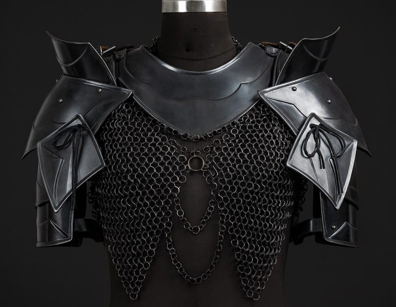 Puede incluir: Una armadura de pecho de metal negro que incluye un gorjal, hombreras articuladas con cordones negros y una cota de malla. La armadura presenta eslabones de cadena decorativos que cuelgan en la parte frontal, asegurados con remaches y placas.