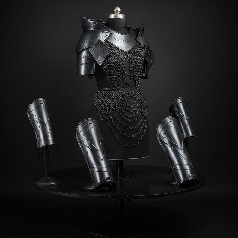 Puede incluir: Un conjunto de armadura negra que incluye una pieza de pecho con cota de malla integrada, hombreras y cuatro grebas a juego. La placa pectoral muestra un dise&ntilde;o en capas con cadenas colgantes, mientras que las grebas presentan una superficie lisa y curva con detalles en forma de V y remaches visibles.