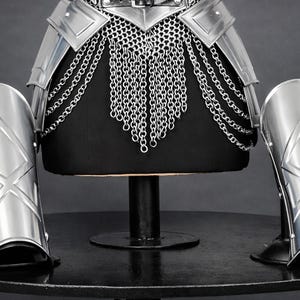 K&ouml;nnte beinhalten: Ein Set aus silberfarbenen Metallr&uuml;stungsteilen, bestehend aus einem H&uuml;ftg&uuml;rtel mit herabh&auml;ngenden Kettengeflechten und vier passenden, gebogenen Beinschienen oder Armschienen. Jedes Teil zeigt ein erhabenes, rautenf&ouml;rmiges Emblem.