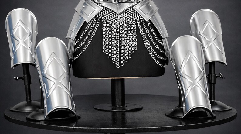 K&ouml;nnte beinhalten: Ein Set aus silberfarbenen Metallr&uuml;stungsteilen, bestehend aus einem H&uuml;ftg&uuml;rtel mit herabh&auml;ngenden Kettengeflechten und vier passenden, gebogenen Beinschienen oder Armschienen. Jedes Teil zeigt ein erhabenes, rautenf&ouml;rmiges Emblem.