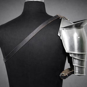 May include: A polished metal shoulder pauldron, part of a suit of armor, is displayed on a black mannequin. The pauldron has a curved design with layered segments for arm movement. A leather strap is attached to the pauldron and crosses the mannequin's back.
