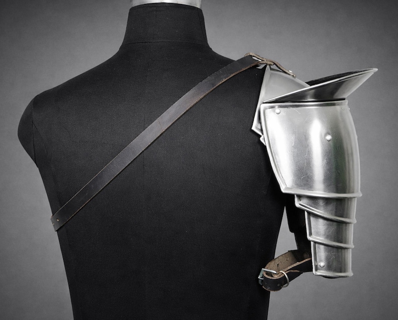 May include: A polished metal shoulder pauldron, part of a suit of armor, is displayed on a black mannequin. The pauldron has a curved design with layered segments for arm movement. A leather strap is attached to the pauldron and crosses the mannequin's back.